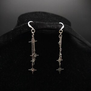 May include: Silver-toned dangle earrings featuring a celestial design. Each earring has a curved hook, a small chain, and multiple star-shaped charms. The earrings are displayed against a black background.
