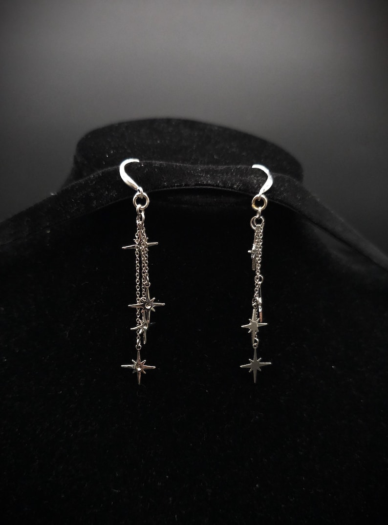 May include: Silver-toned dangle earrings featuring a celestial design. Each earring has a curved hook, a small chain, and multiple star-shaped charms. The earrings are displayed against a black background.