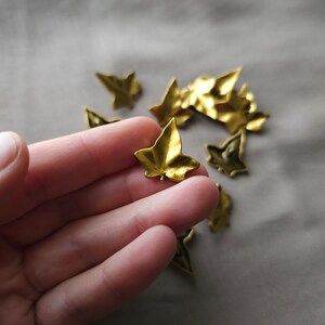 May include: A hand holding a gold-colored metal leaf charm. The leaf is a simple, five-pointed design. There are several other similar leaf charms scattered on a light brown surface.