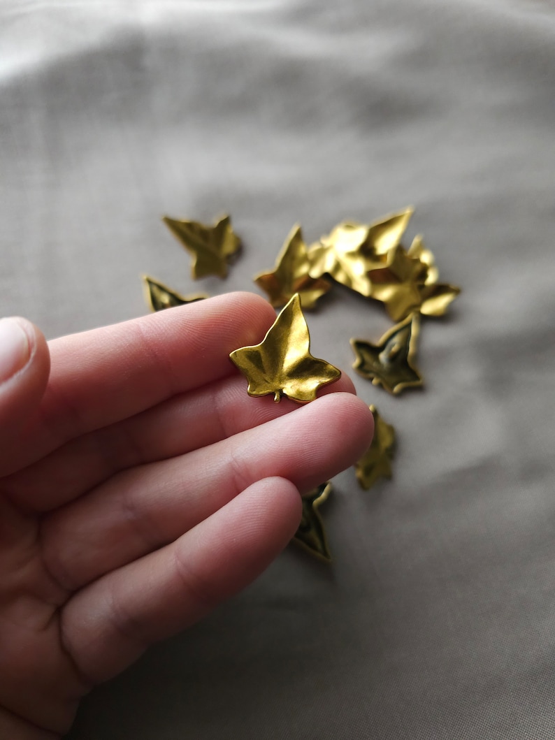 May include: A hand holding a gold-colored metal leaf charm. The leaf is a simple, five-pointed design. There are several other similar leaf charms scattered on a light brown surface.