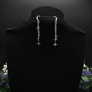 May include: Silver-toned dangle earrings with a celestial theme. Each earring features a delicate chain with star and starburst charms. The earrings are displayed on a black mannequin head against a dark background, highlighting their intricate design and sparkle.