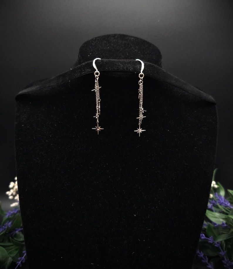 May include: Silver-toned dangle earrings with a celestial theme. Each earring features a delicate chain with star and starburst charms. The earrings are displayed on a black mannequin head against a dark background, highlighting their intricate design and sparkle.