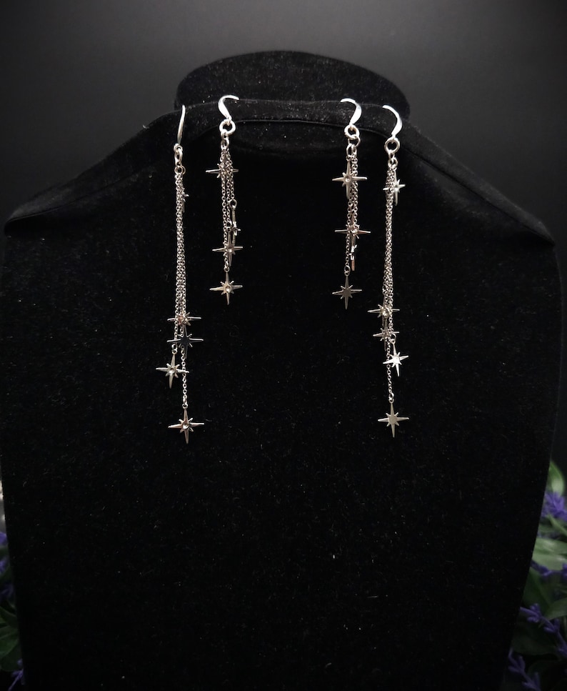 May include: Silver-toned dangle earrings featuring a series of star charms suspended from delicate chains. The earrings have a hook closure and are displayed on a black mannequin head. The stars are evenly spaced along the chains.