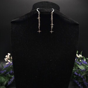 May include: Silver-toned dangle earrings with a star and chain design. The earrings are displayed on a black velvet bust, with a dark background. The earrings have a hook closure and feature multiple star charms.