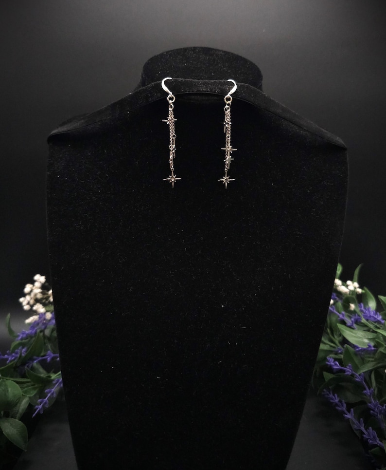 May include: Silver-toned dangle earrings with a star and chain design. The earrings are displayed on a black velvet bust, with a dark background. The earrings have a hook closure and feature multiple star charms.