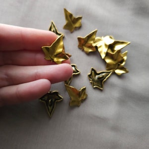 May include: A collection of gold-colored metal ivy leaf charms. The leaves are scattered on a light beige fabric surface.