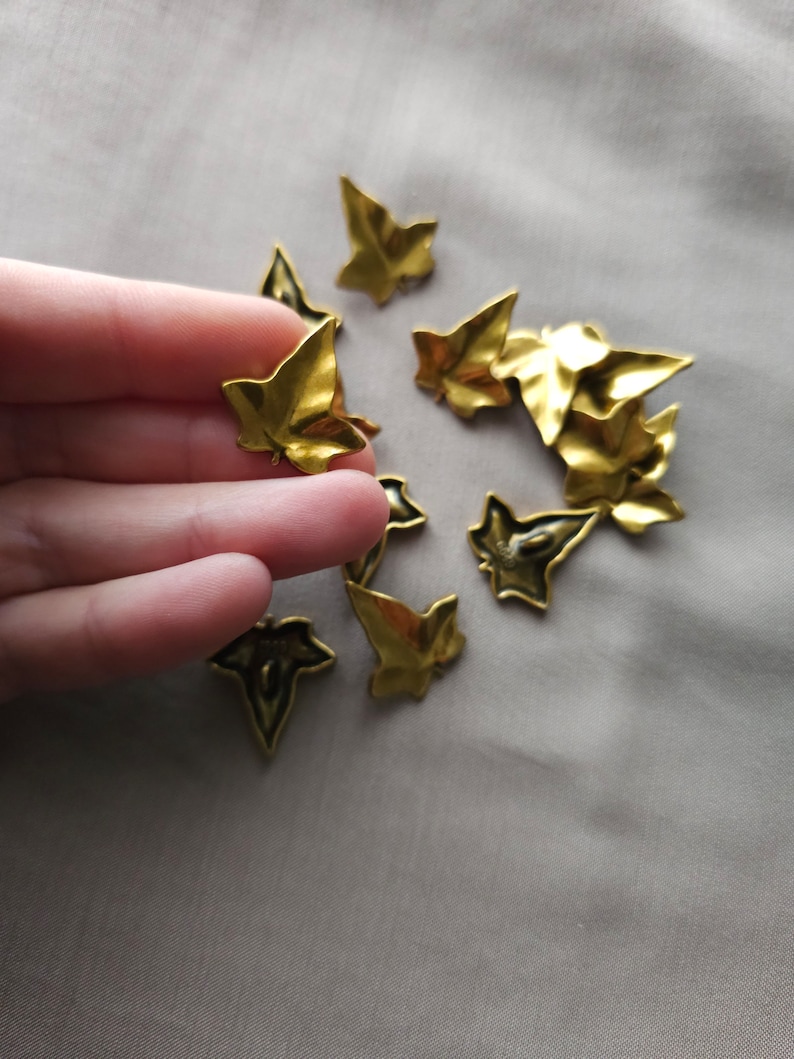 May include: A collection of gold-colored metal ivy leaf charms. The leaves are scattered on a light beige fabric surface.