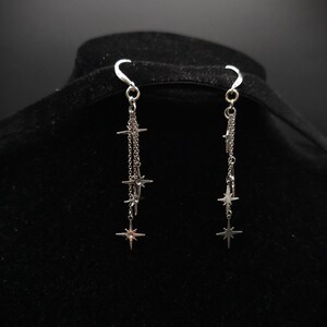 May include: Silver dangle earrings featuring a star design. Each earring has a hook and multiple chains with star charms of varying sizes. The earrings are displayed against a black background.