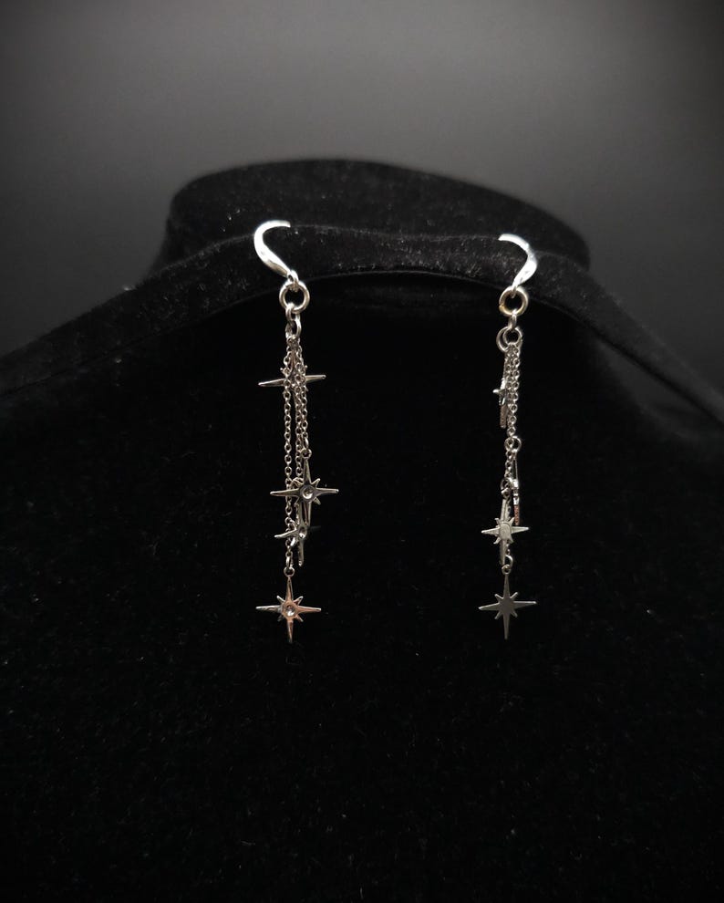 May include: Silver dangle earrings featuring a star design. Each earring has a hook and multiple chains with star charms of varying sizes. The earrings are displayed against a black background.