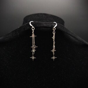 May include: A pair of silver-toned dangle earrings. Each earring features a chain with multiple star-shaped charms of varying sizes. The earrings are suspended from a curved hook and are displayed against a dark background.