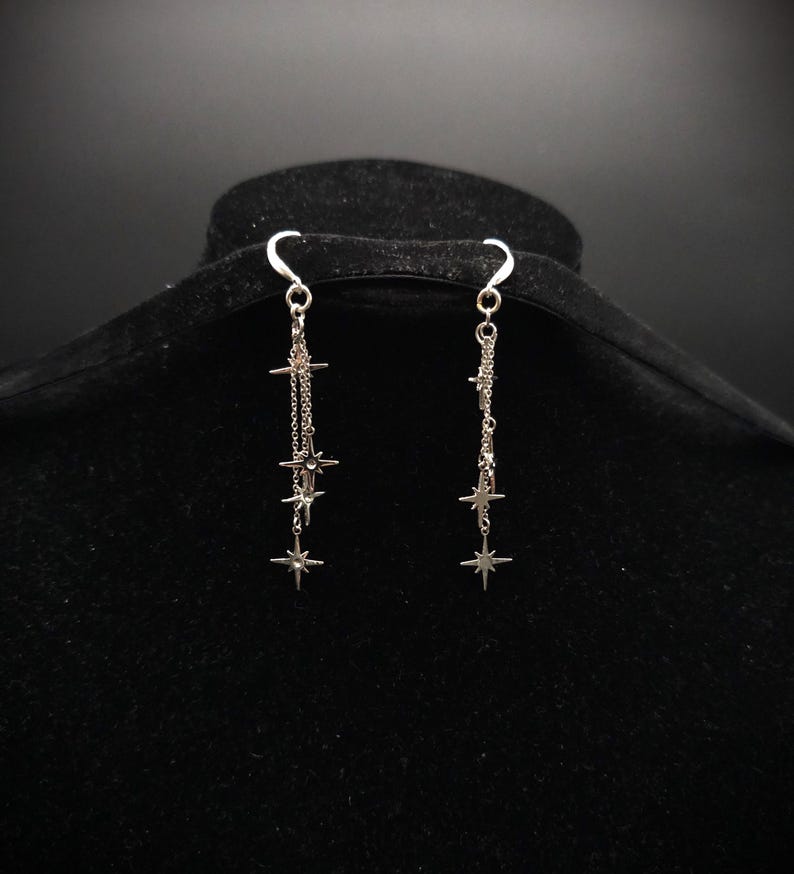 May include: A pair of silver-toned dangle earrings. Each earring features a chain with multiple star-shaped charms of varying sizes. The earrings are suspended from a curved hook and are displayed against a dark background.