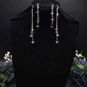 May include: Silver-toned dangle earrings featuring a cascade of star charms. The earrings hang from delicate chains, with multiple stars of varying sizes. The earrings are displayed on a black mannequin head against a dark background.