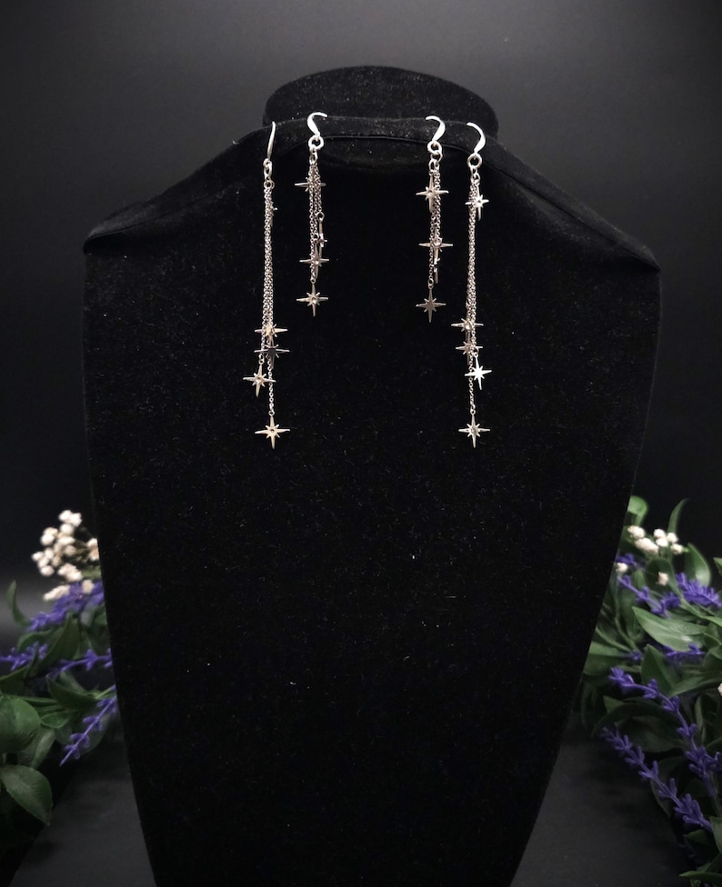May include: Silver-toned dangle earrings featuring a cascade of star charms. The earrings hang from delicate chains, with multiple stars of varying sizes. The earrings are displayed on a black mannequin head against a dark background.