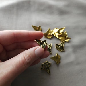 May include: A collection of gold-colored metal ivy leaf embellishments. The leaves are scattered on a light beige surface. One leaf is being held in a hand.