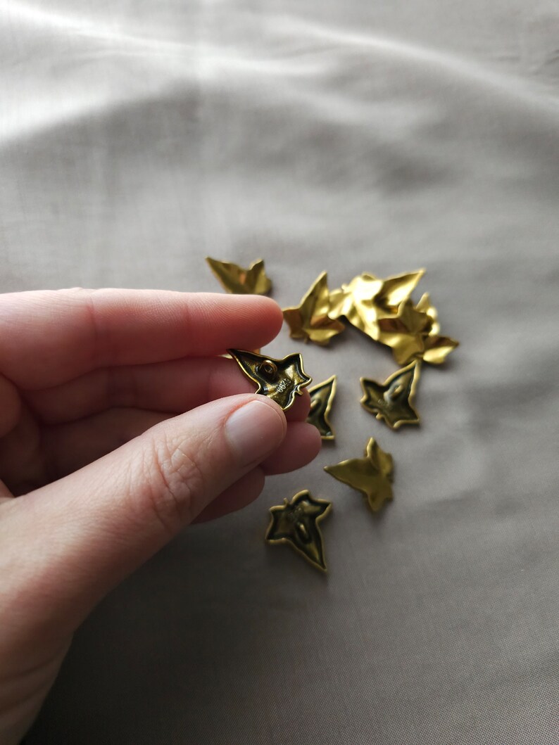 May include: A collection of gold-colored metal ivy leaf embellishments. The leaves are scattered on a light beige surface. One leaf is being held in a hand.