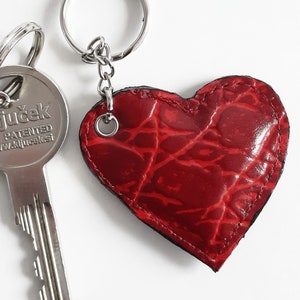Può includere: Un portachiavi a forma di cuore in pelle rossa con un motivo a coccodrilo. Il portachiavi è attaccato a un anello e una catena argentati.