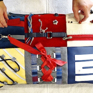 Puede incluir: Una almohada sensorial multicolor con varias texturas y objetos para la estimulación táctil. Presenta una cinta roja, una cremallera, una llave, una hebilla, un botón, un cordón trenzado y una variedad de otras texturas.