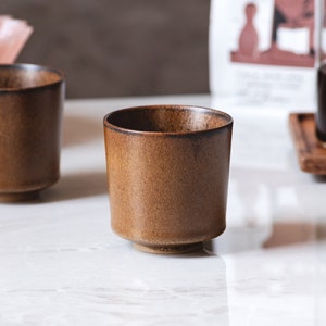May include: Two brown ceramic mugs on a white marble surface. The mugs are slightly different shades of brown and have a rounded shape.
