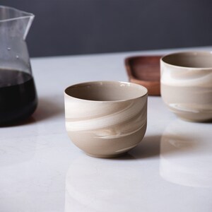 May include: Two beige and white swirl-patterned ceramic cups sit on a white surface, near a glass carafe filled with dark liquid and a wooden tray. The cups have a rounded shape and a smooth finish, ideal for serving beverages.