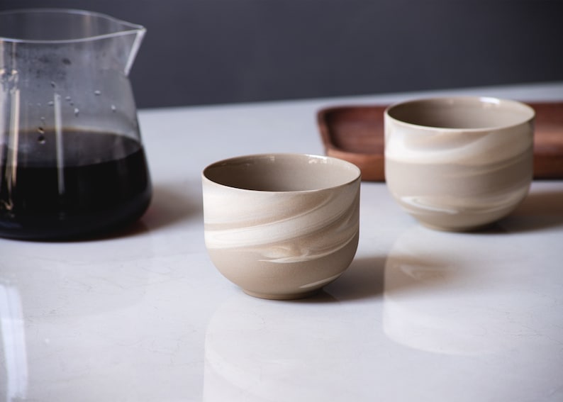 May include: Two beige and white swirl-patterned ceramic cups sit on a white surface, near a glass carafe filled with dark liquid and a wooden tray. The cups have a rounded shape and a smooth finish, ideal for serving beverages.