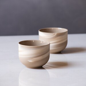 May include: Two ceramic bowls with a beige and white swirl pattern. The bowls are cylindrical with a rounded base and a smooth, glossy finish. The bowls are displayed on a white surface against a gray background.