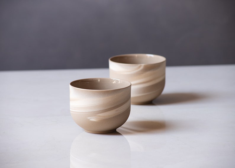 May include: Two ceramic bowls with a beige and white swirl pattern. The bowls are cylindrical with a rounded base and a smooth, glossy finish. The bowls are displayed on a white surface against a gray background.