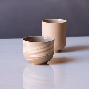 May include: Two ceramic vessels in neutral tones. One is a rounded bowl with a swirling, marbled pattern in shades of beige and cream. The other is a tall, cylindrical vase in a solid, light beige color, with a rounded base.