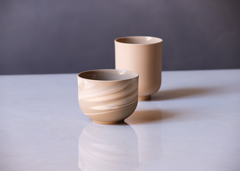 May include: Two ceramic vessels in neutral tones. One is a rounded bowl with a swirling, marbled pattern in shades of beige and cream. The other is a tall, cylindrical vase in a solid, light beige color, with a rounded base.