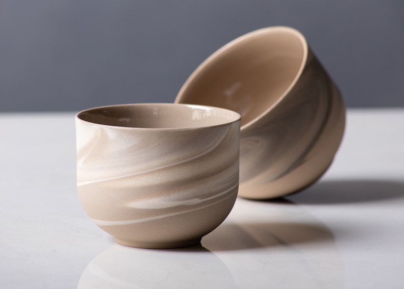 Front view of two minimalist, round, stoneware cup in brown with white swirls on white marble. One is on it's side.