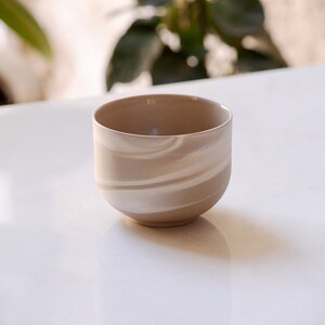 Front view of a minimalist, round, stoneware cup in brown with white swirls on white marble with plants in background.