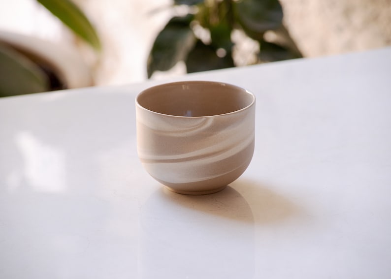 Front view of a minimalist, round, stoneware cup in brown with white swirls on white marble with plants in background.