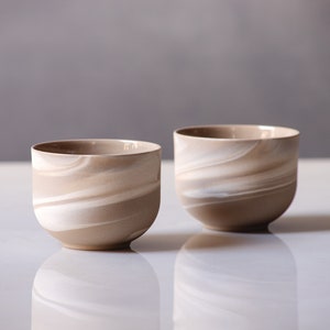 May include: Two ceramic cups with a swirling pattern of beige and white. The cups have a rounded shape and a smooth, glossy finish. The cups are likely for drinking beverages.