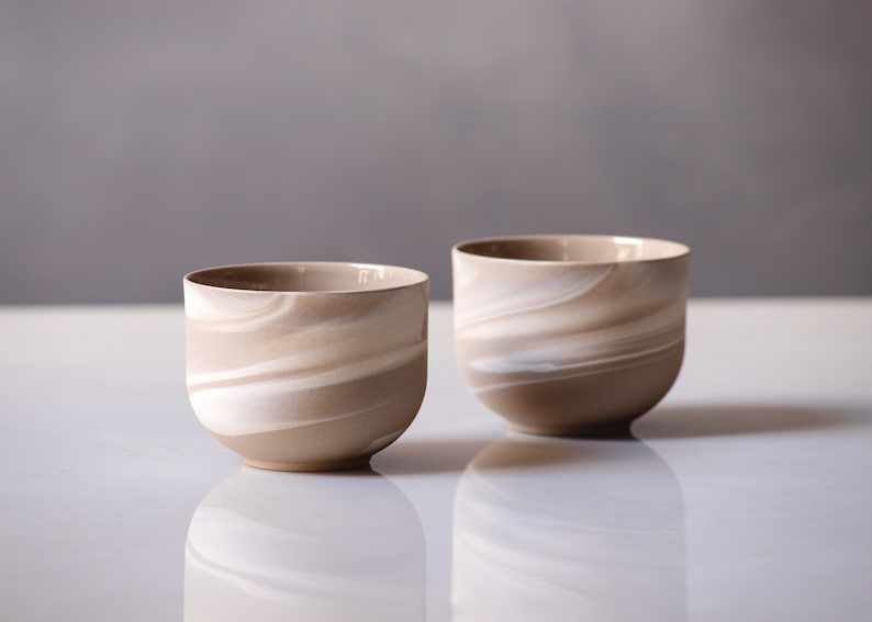 May include: Two ceramic cups with a swirling pattern of beige and white. The cups have a rounded shape and a smooth, glossy finish. The cups are likely for drinking beverages.