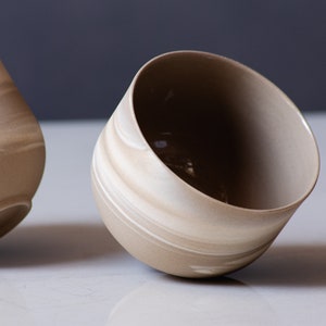 May include: Two beige ceramic bowls with a marbled design. The bowls have a rounded shape with a wide opening. The interior of the bowls is a darker shade of beige. The bowls are tilted slightly, showcasing their design.
