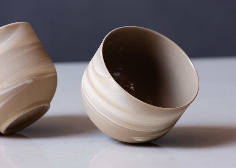 May include: Two beige ceramic bowls with a marbled design. The bowls have a rounded shape with a wide opening. The interior of the bowls is a darker shade of beige. The bowls are tilted slightly, showcasing their design.