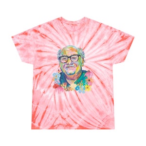 May include: A tie-dye t-shirt with a colorful portrait of a man wearing glasses and surrounded by flowers. The shirt is a light pink color with a swirl pattern.
