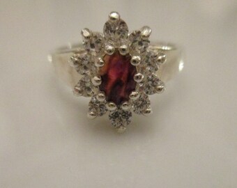 Puede incluir: Un anillo de plata con una gran piedra preciosa ovalada, de color púrpura iridiscente, rodeada de un grupo de piedras preciosas más pequeñas y transparentes.