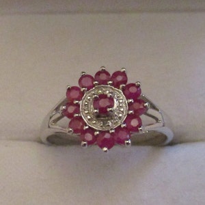 Puede incluir: Un anillo de plata con un grupo de rubíes rojos engastados en forma de flor. El centro de la flor tiene una piedra más pequeña y transparente.