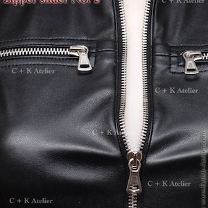 May include: Close-up of a black leather jacket zipper with a silver slider. The zipper is partially open, revealing the white lining of the jacket. The text "C+K Atelier" is visible on the jacket.