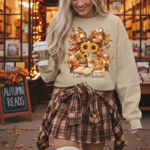 Könnte beinhalten: Beiges Sweatshirt mit einem Herbst-Motiv mit Sonnenblumen, Kürbissen und einer karierten Schleife. Der Text "I am an Autumn" ist darunter gedruckt. Die Person trägt einen karierten Rock und hält eine Kaffeetasse.