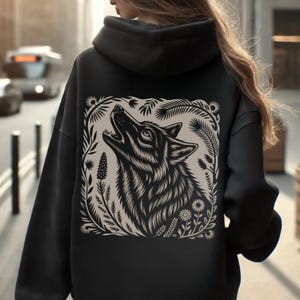 May include: Black hoodie with a detailed illustration of a howling wolf framed by floral and foliage elements. The design uses grey and beige tones, contrasting against the dark fabric. The hoodie has a hood and long sleeves.
