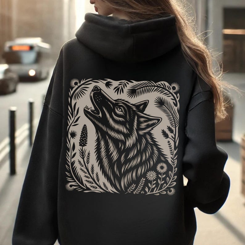 Model wearing a hoodie with a large linocut-style howling wolf illustration on the back, surrounded by botanical elements. Design shown in black, forest green, maroon, and dark chocolate color options.