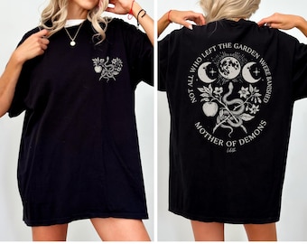 May include: Black t-shirt with a white collar, featuring a front graphic of a snake, apple, and floral design. The back of the shirt displays a circular design with the text "NOT ALL WHO LEFT THE GARDEN WERE BANISHED MOTHER OF DEMONS".