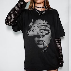 Op de afbeelding: Zwart grafisch T-shirt met een zwart-wit afbeelding van een persoon die rookt. De hand van de persoon bedekt hun ogen, met de woorden "NOT FROM THE RIB" getatoeëerd op de vingers. Het T-shirt heeft korte mouwen. De persoon draagt een choker.