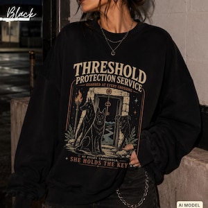 May include: A black long-sleeve sweatshirt featuring a graphic print with the text "THRESHOLD PROTECTION SERVICE GUARDED AT EVERY CROSSING" and "AT EVERY THRESHOLD, SHE HOLDS THE KEY". The design depicts three dogs near a stone doorway with a key.