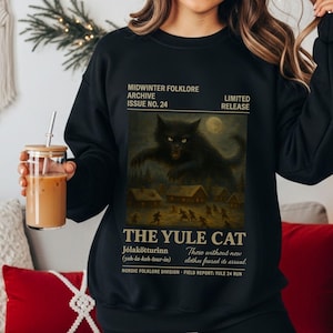 May include: Black sweatshirt featuring a vintage-style illustration of a large black cat with glowing eyes, set against a night scene with a full moon. The text reads "MIDWINTER FOLKLORE ARCHIVE ISSUE NO. 24" and "THE YULE CAT".