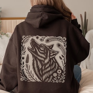 Model wearing a hoodie with a large linocut-style howling wolf illustration on the back, surrounded by botanical elements. Design shown in black, forest green, maroon, and dark chocolate color options.