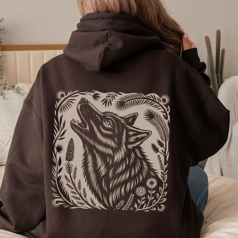 Model wearing a hoodie with a large linocut-style howling wolf illustration on the back, surrounded by botanical elements. Design shown in black, forest green, maroon, and dark chocolate color options.