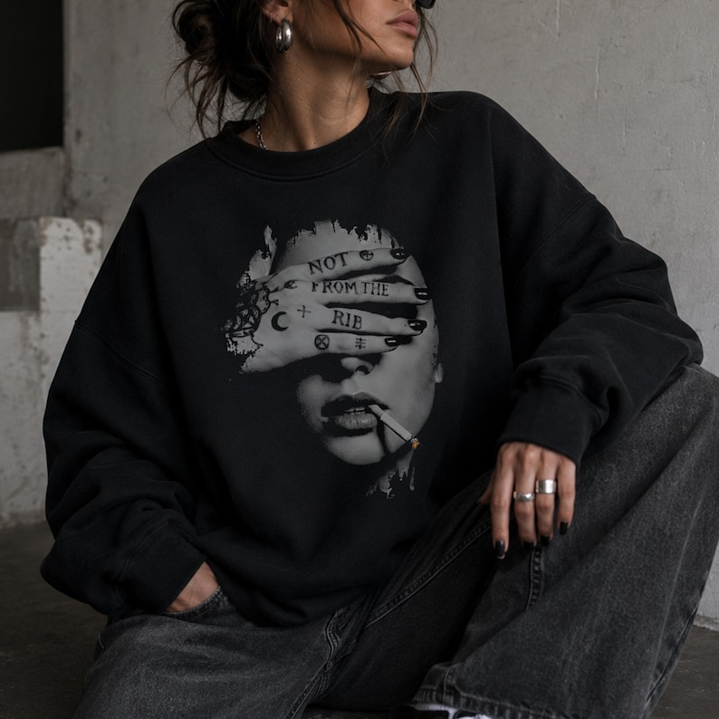 Seated woman modeling a black Gildan 18000 Lilith crewneck sweatshirt with a dark feminine hand-over-face graphic and “Not From the Rib” quote.