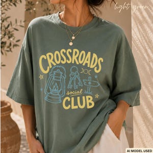 Könnte beinhalten: Ein hellgrünes T-Shirt mit dem gelben Schriftzug "CROSSROADS social CLUB". Die Grafik zeigt eine blaue Laterne, einen Schlüsselbund, ein hölzernes Wegweiser-Schild und kleine Sternsymbole. Das T-Shirt hat einen Rundhalsausschnitt.